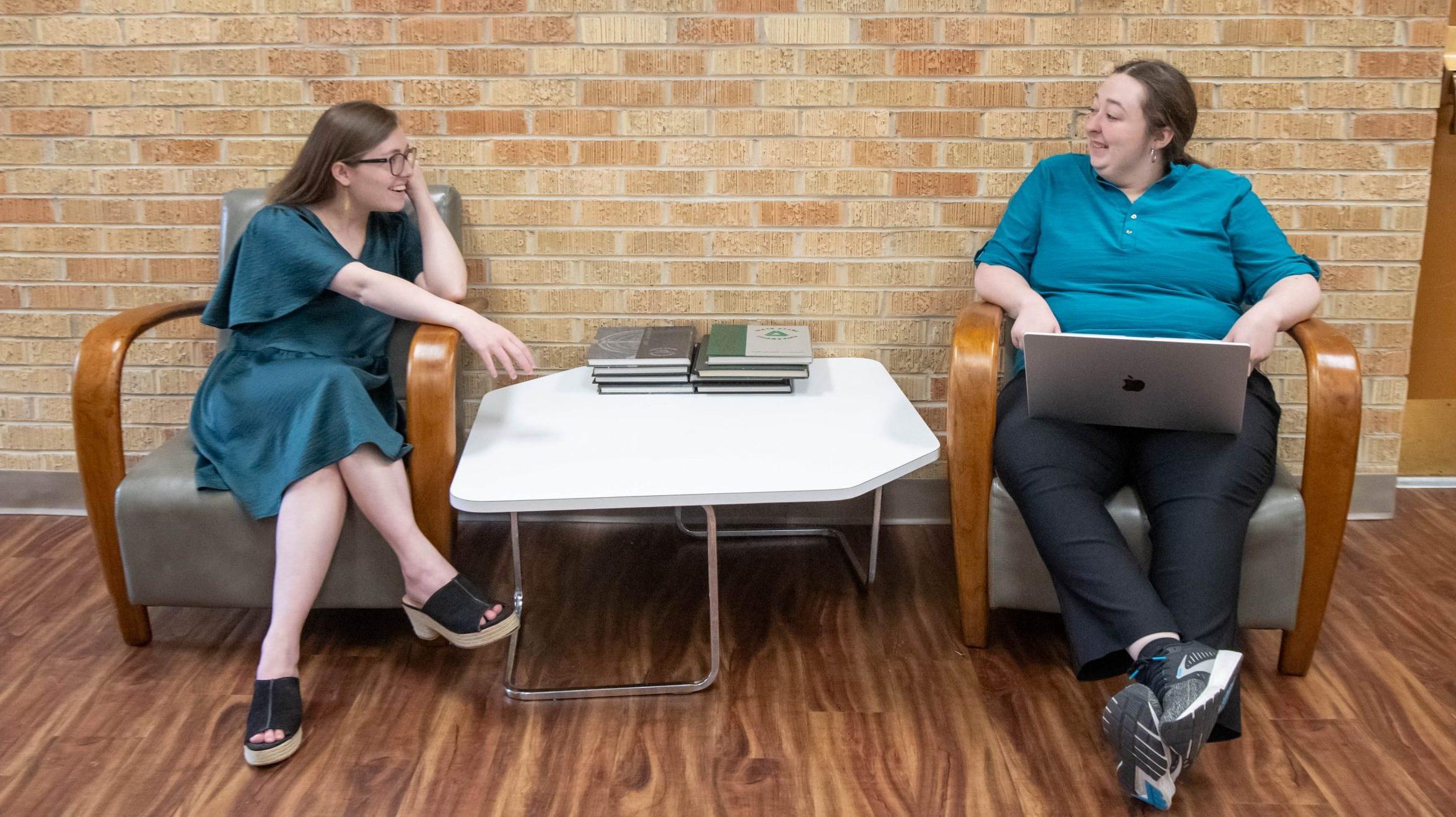 Two students having a conversation in a sitting area, one with a laptop.